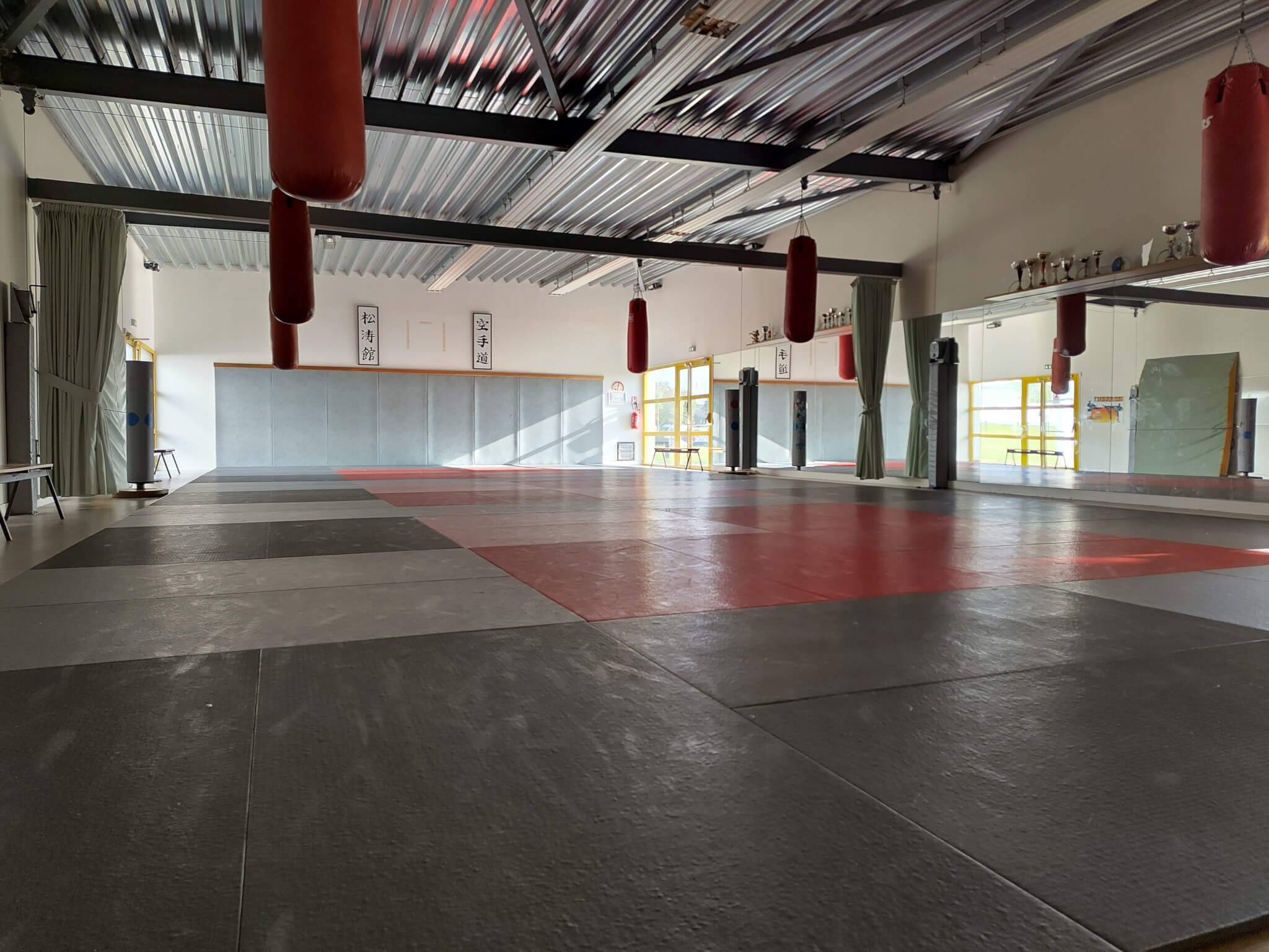 Intérieur du dojo Ange Jacquier à Thouaré sur Loire. Le sol est couvert de tatamis et des sacs de frappe sont suspendus au plafond. Un des murs est couvert de miroirs